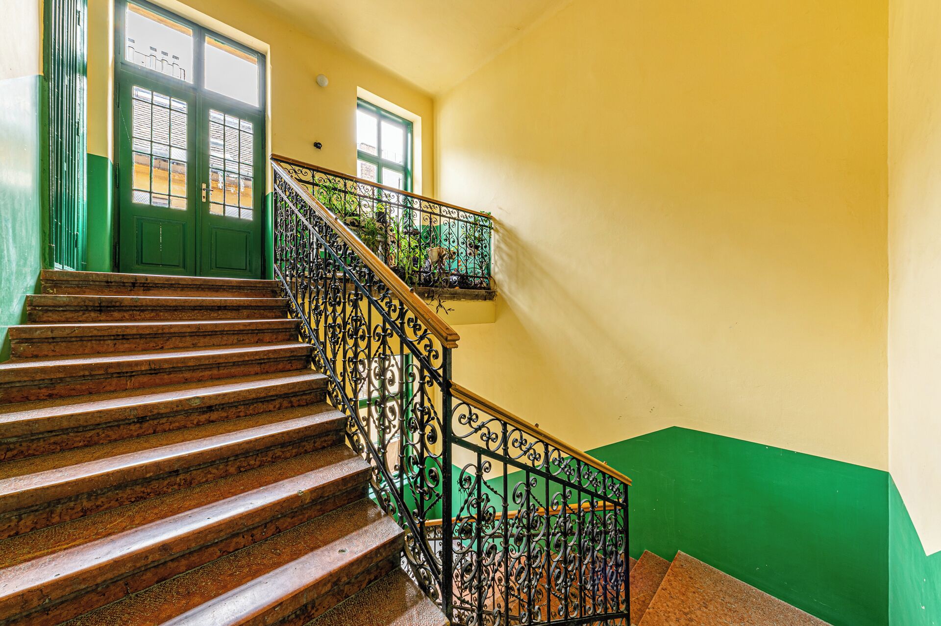 A staircase with wooden steps and black iron railings, leading to a landing with a green wall and a window.