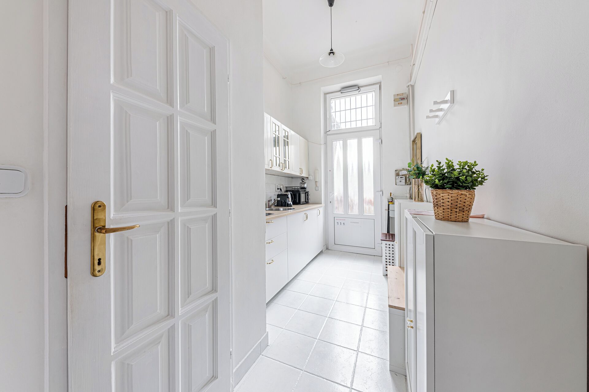 A narrow hallway with white walls and doors, leading to a kitchen area with white cabinets and a window.