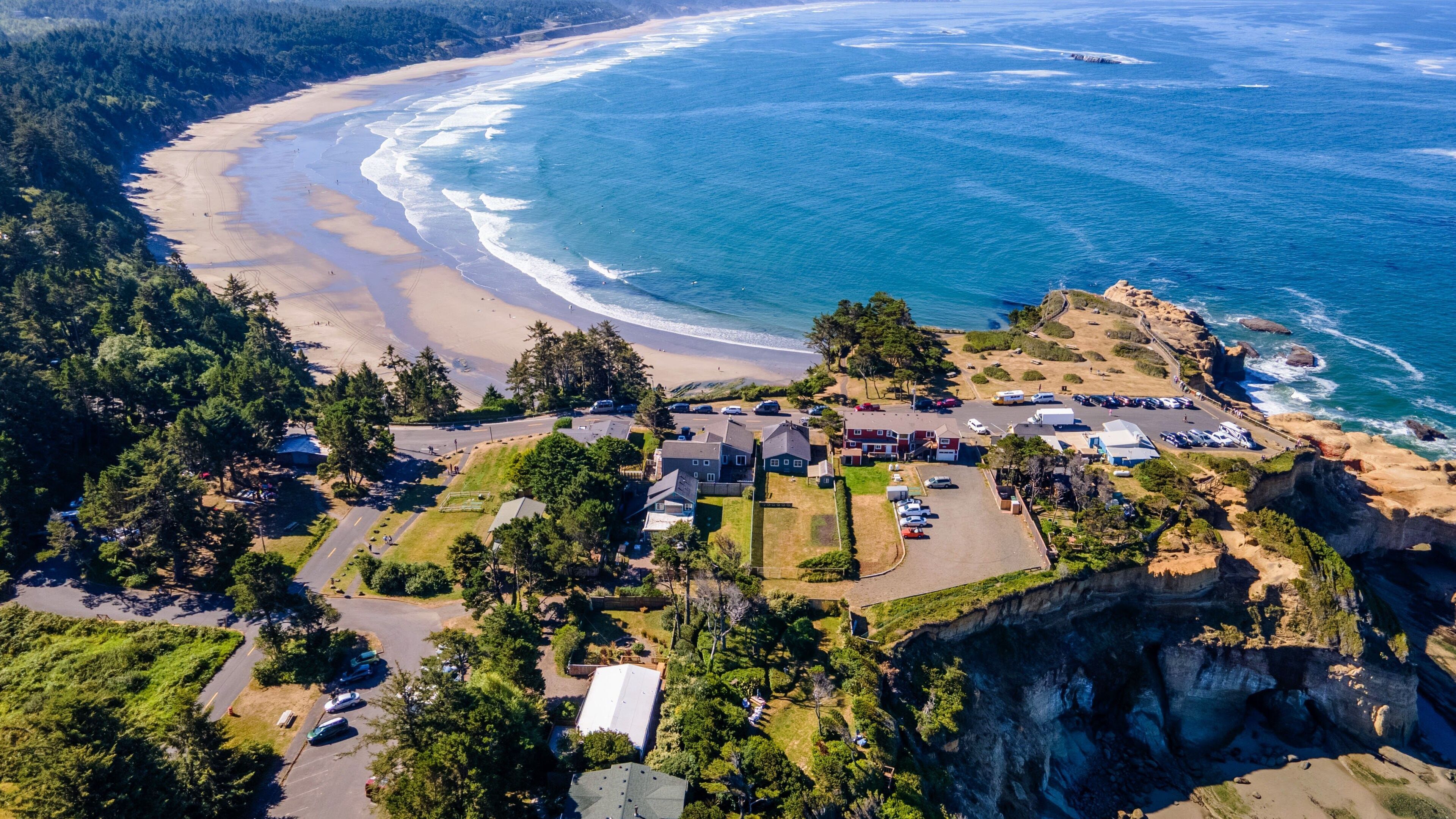 An aerial view of a coastal property with a beach, rocky cliffs, and a parking area.