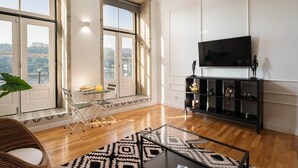 A spacious living room with a large window, a flat-screen TV, a glass coffee table, and a black entertainment unit.