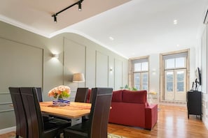 A dining area with a table, chairs, a vase of flowers, a red sofa, and a window with a view.