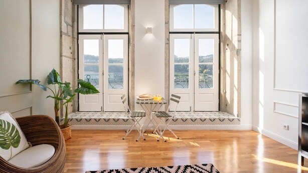 A room with a wooden floor, a dining table with chairs, a wicker chair, a potted plant, and large windows with a view.