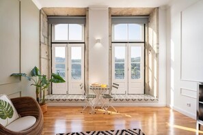A room with a wooden floor, a dining table with chairs, a wicker chair, a potted plant, and large windows with a view. - Luxury stay premium Douro river & sunset views (porto)