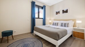 Et moderne hotelværelse med en seng, to sengeborde, en lille skammel og et vindue med gardiner.