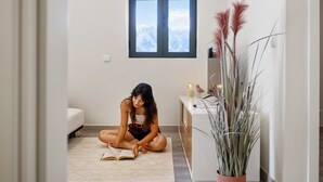 Eine Person sitzt auf dem Boden und liest ein Buch in einem Raum mit Fenster, weißem Schreibtisch und einer Topfpflanze.