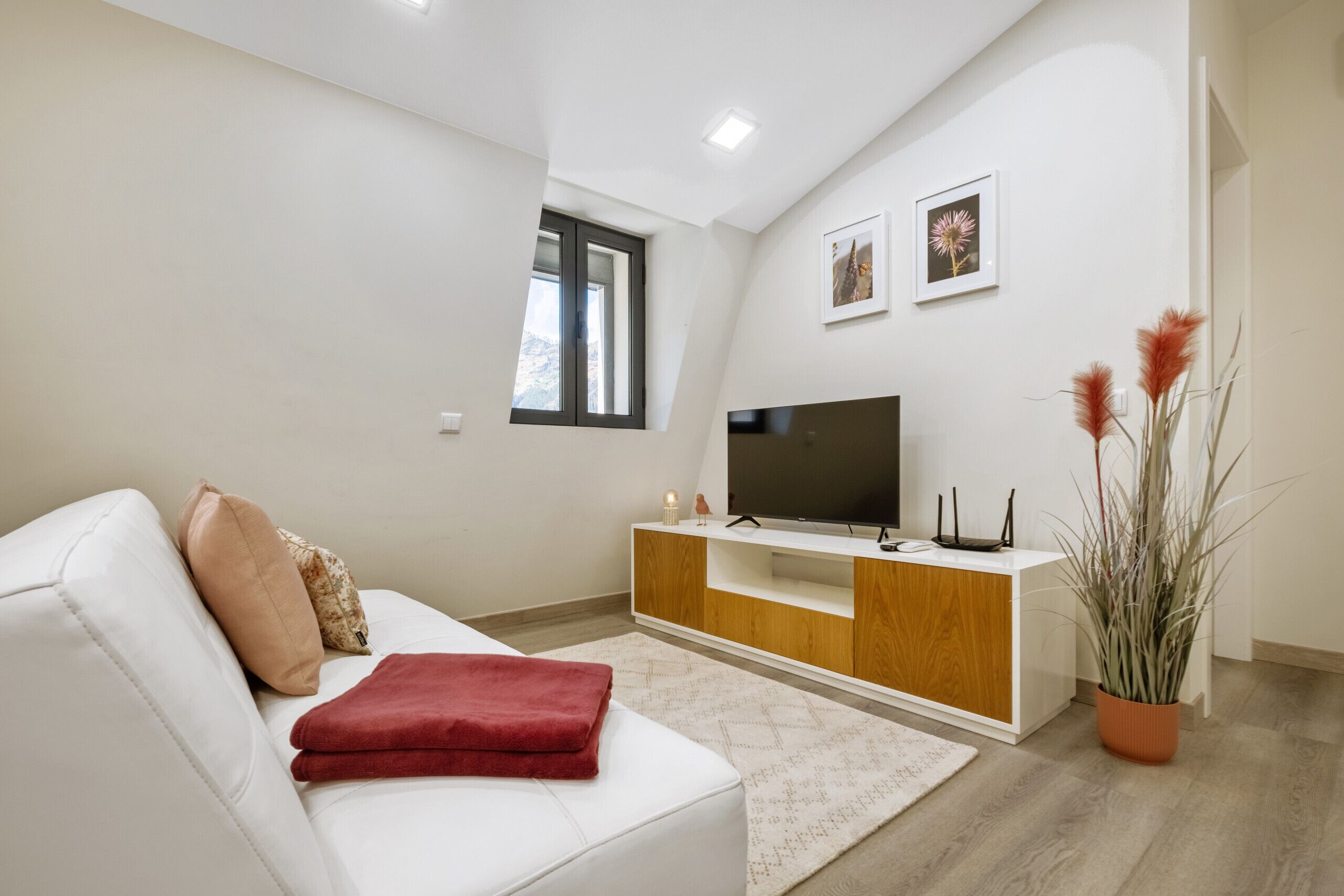 A modern living room with a white sofa, a flat-screen TV on a wooden stand, a small window, and a potted plant.