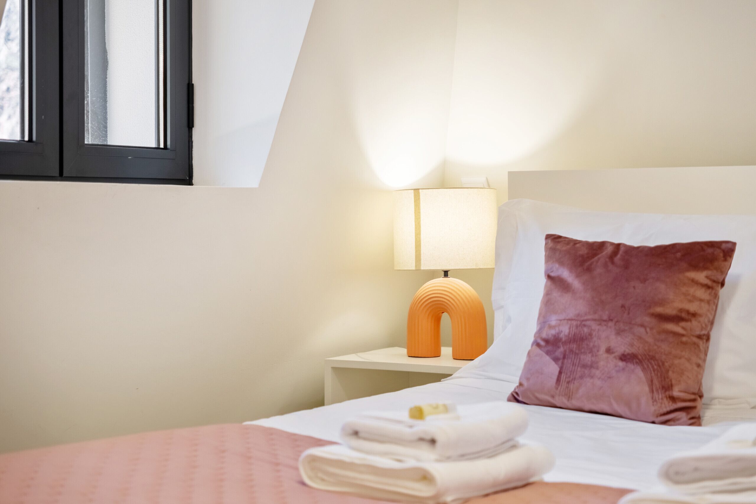 A neatly made bed with folded towels, a bedside lamp, and a window with blinds.