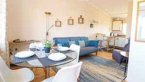 Un salon moderne avec une table à manger en verre, des chaises blanches, un canapé bleu et un coin cuisine à l’arrière-plan.