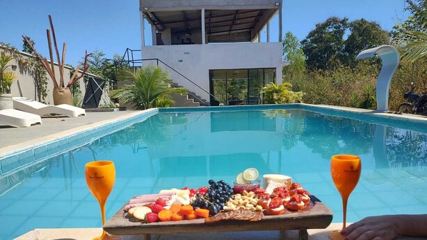 Vista de la piscina con una tabla de frutas y quesos, sillas de descanso y un edificio moderno al fondo.