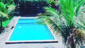Una piscina rectangular con borde de baldosas, rodeada de abundante vegetación y un edificio con techo rojo al fondo.