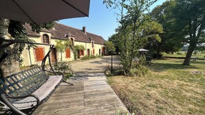 A wooden deck with a bench, a pathway leading to a building with red shutters, and a grassy area with trees. - Entire vacation home in Burgundy style: Longère des Daubénys (Precy-sur-Vrin)