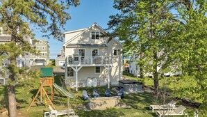 A large two-story house with a balcony, a swing set, and a backyard with lawn chairs and a fire pit.