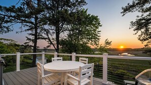 A wooden deck with a white table and chairs, overlooking a sunset over a river.