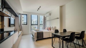 Moderni hotellihuone, jossa on pieni keittiö, ruokailutila pöytineen ja tuoleineen, sängystä avautuva kaunis näkymä ja suuret ikkunat.