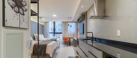 Een moderne hotelkamer met kitchenette, een bed met twee kussens, een kleine tafel met stoel en een groot raam.