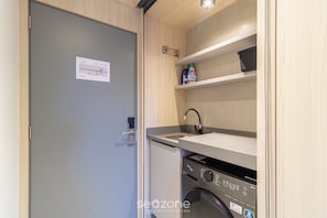 Cocina compacta con lavadora integrada, una pequeña encimera y una estantería con productos de limpieza.