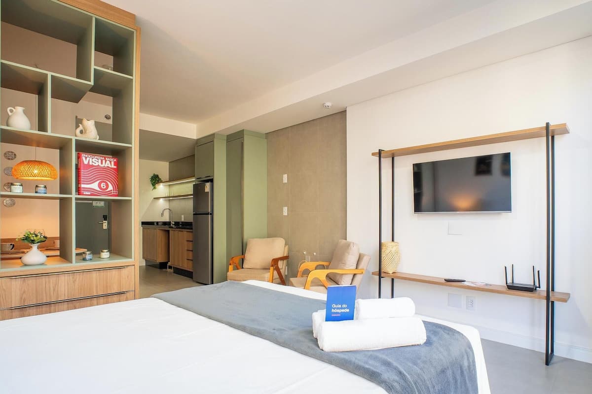A modern hotel room with a bed, a TV mounted on the wall, a bookshelf, and a kitchenette.