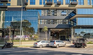 A modern building with a glass facade, a parking area with cars, and a motorcycle. - Modern and Fully Equipped Apartments Just Minutes From Lagoa da Conceicao and the Beach - VTD (Florianópolis)