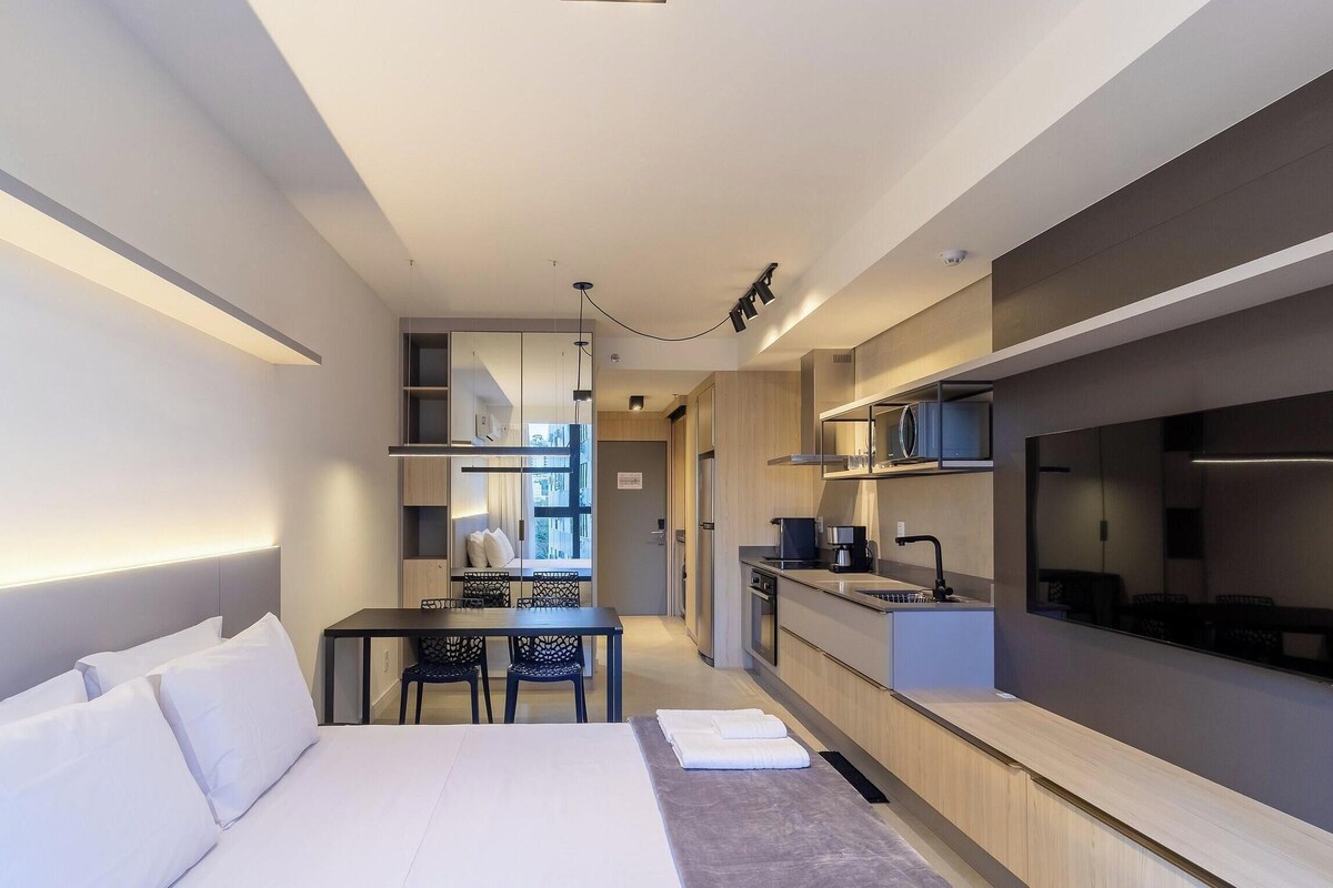 A modern hotel room with a bed, a small dining table, a kitchenette, and a TV.