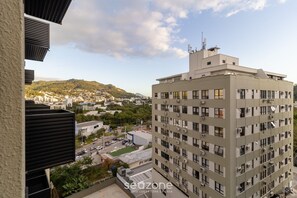 A multi-story building with numerous windows and air conditioning units, a flat roof, and a view of a cityscape with greenery and hills in the background. - Modern and Fully Equipped Apartments Just Minutes From Lagoa da Conceicao and the Beach - VTD (Florianópolis)