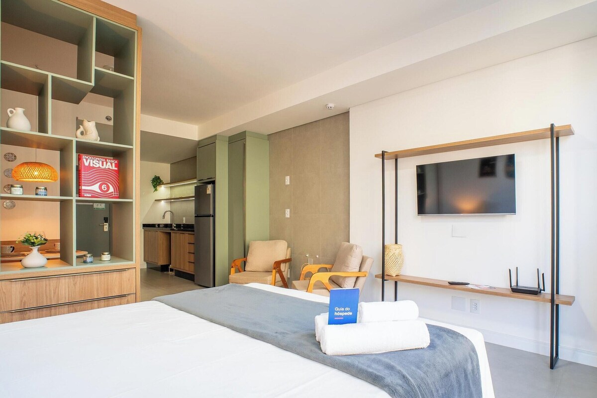 A modern hotel room with a bed, a TV mounted on the wall, a bookshelf, and a kitchenette.