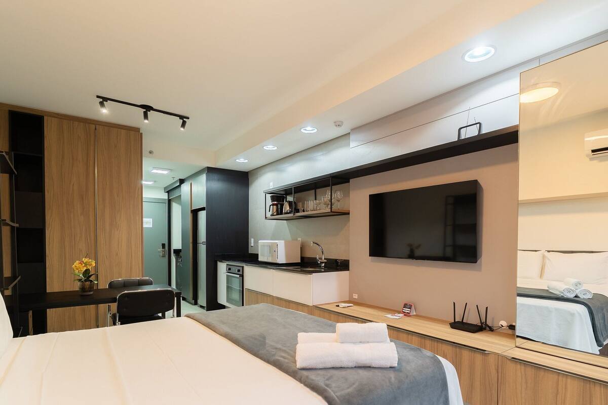 A modern hotel room with a bed, a desk, a kitchenette, and a large mirror.