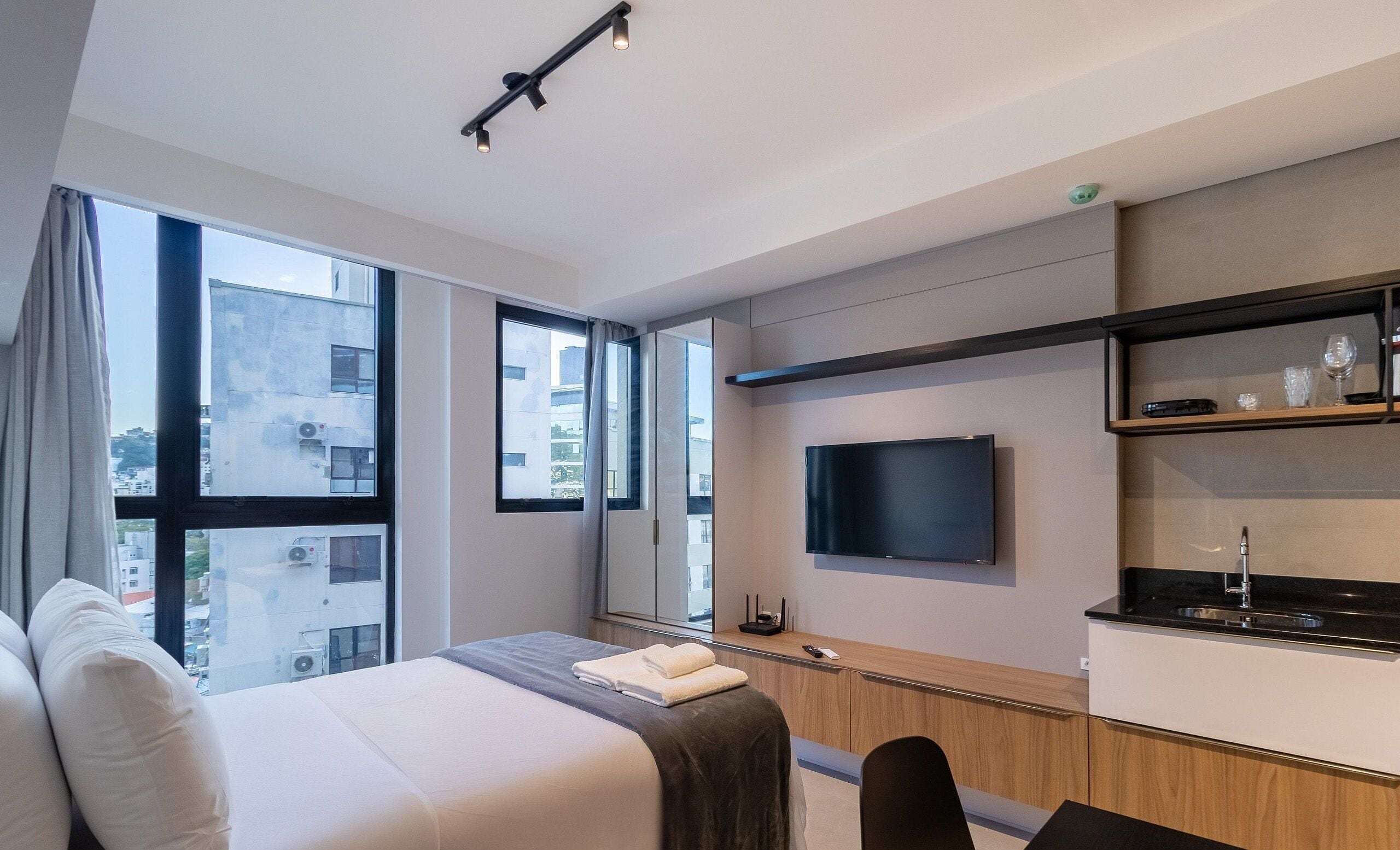 Una habitación de hotel moderna con una cama grande, un televisor montado en la pared, una pequeña kitchenette y ventanas amplias con vistas a la ciudad.
