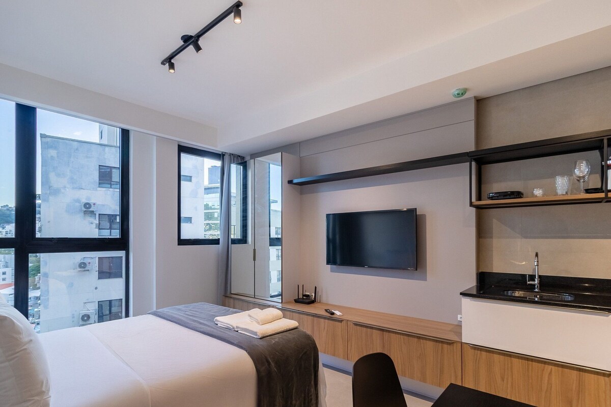 A modern hotel room with a large bed, a TV mounted on the wall, a small kitchenette, and large windows offering a city view.