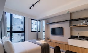 Una habitación de hotel moderna con una cama grande, un televisor montado en la pared, una pequeña kitchenette y ventanas amplias con vistas a la ciudad.