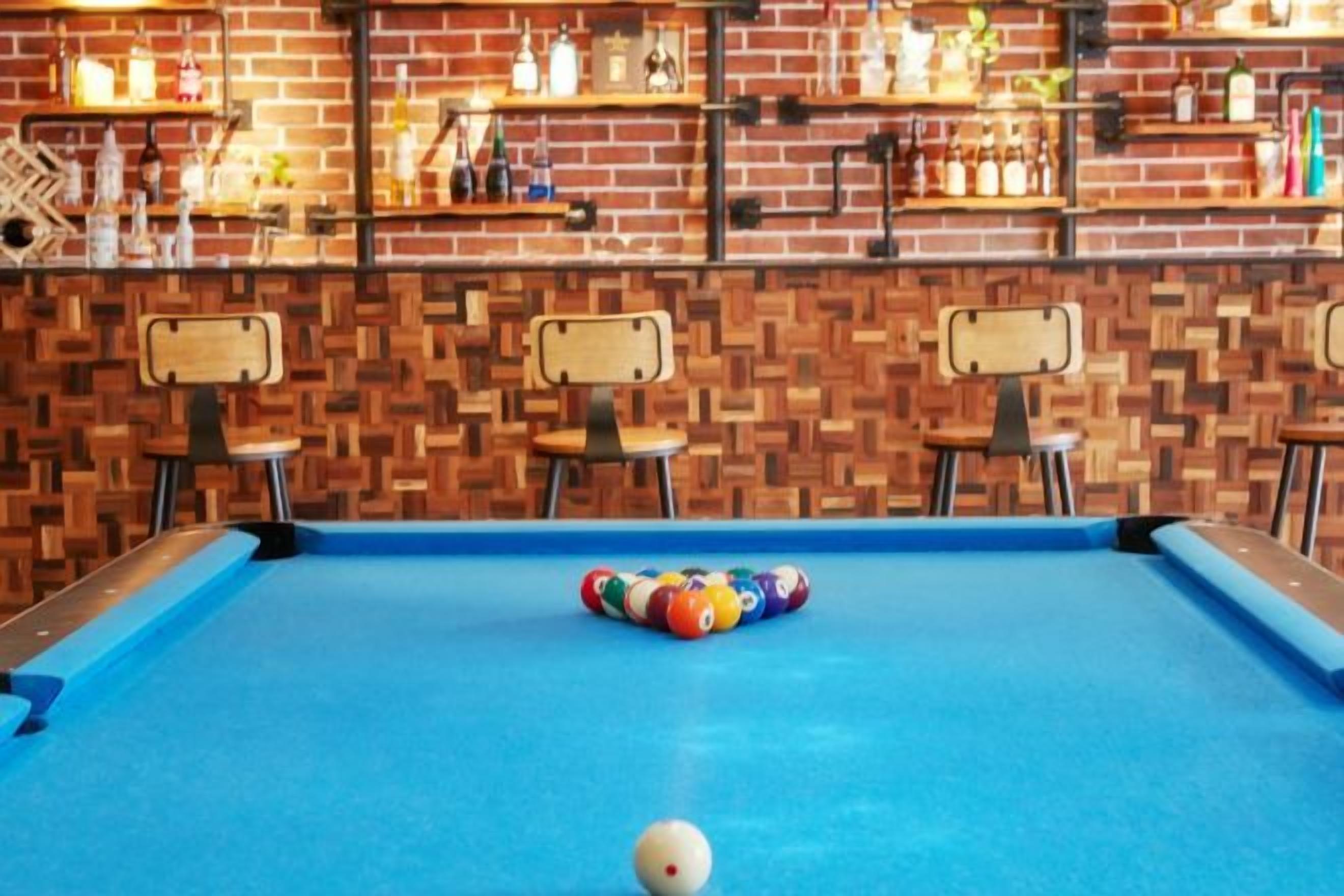 A pool table with a ball, surrounded by chairs, in a room with a brick wall and shelves with bottles.