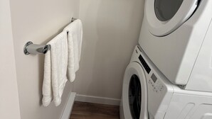 A laundry area with a stacked washer and dryer, a wooden floor, and a towel rack with towels. - New suite in a great location. Central for shopping, restaurants and commuting. (Regina)