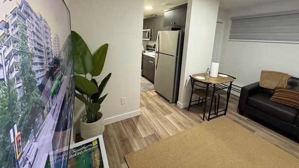 A modern living room with a large flat-screen TV, a dining area with a small table and chairs, a kitchen with a refrigerator and microwave, and a potted plant. - New suite in a great location. Central for shopping, restaurants and commuting. (Regina)