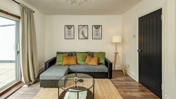 A modern living room with a sofa, a glass coffee table, and a chandelier. There are three framed artworks on the wall and a door to the right.