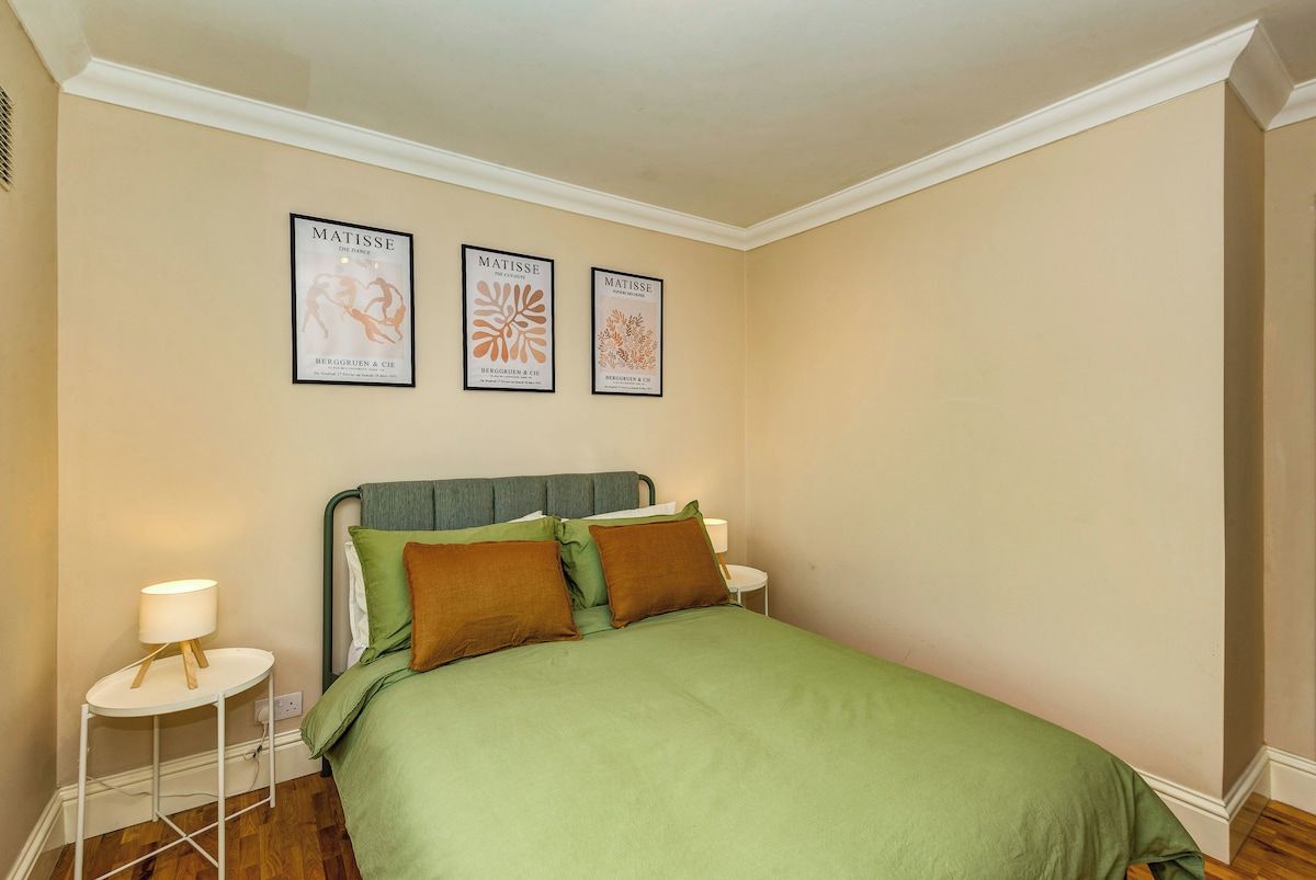 A bedroom with a bed, green bedding, two pillows, a nightstand with a lamp, and three framed artworks on the wall.