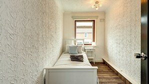 A narrow hallway with textured white walls, a wooden floor, a bed with white bedding and a folded blanket, a desk with a chair, and a window with blinds.