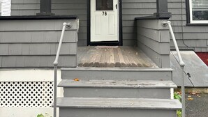 A gray house with a white door marked with the number 76, a star decoration, and a covered porch with steps leading up to the entrance. - Coastal Charm  – Walk to Beach or Train! (Swampscott)