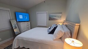 A bedroom with a bed, a TV, a lamp, and a painting on the wall. - Tybee Beach Cottage, Steps from all the local fun and a quick walk to the beach (Tybee Island)