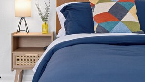 A neatly made bed with a blue comforter and a colorful geometric pillow, a bedside table with a lamp and a vase with a plant, and a wooden headboard.