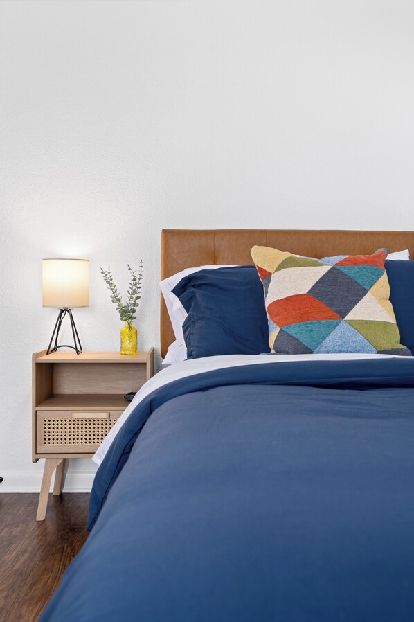 Una cama bien hecha con una colcha azul y un cojín geométrico de colores, una mesita de noche con una lámpara y un florero con una planta, y un cabecero de madera.