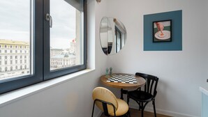 Une pièce avec une fenêtre, une table ronde sur laquelle est posé un jeu d’échecs, deux chaises, un miroir et un tableau encadré accroché au mur.