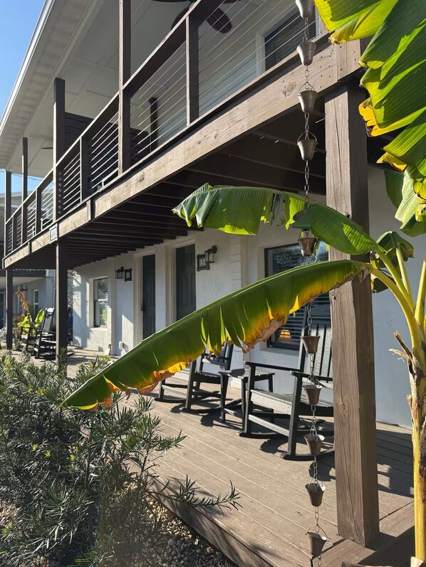 Een houten terras met buitenmeubilair, een hangende plantenbak met een bananenplant, en een gebouw met een balkon.