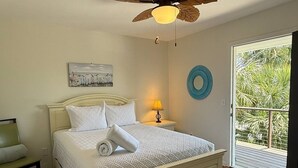 Ein Schlafzimmer mit Bett, Deckenventilator, Nachttisch, Lampe, Stuhl, einem gemusterten Teppich und Blick ins Grüne durch eine Glastür.