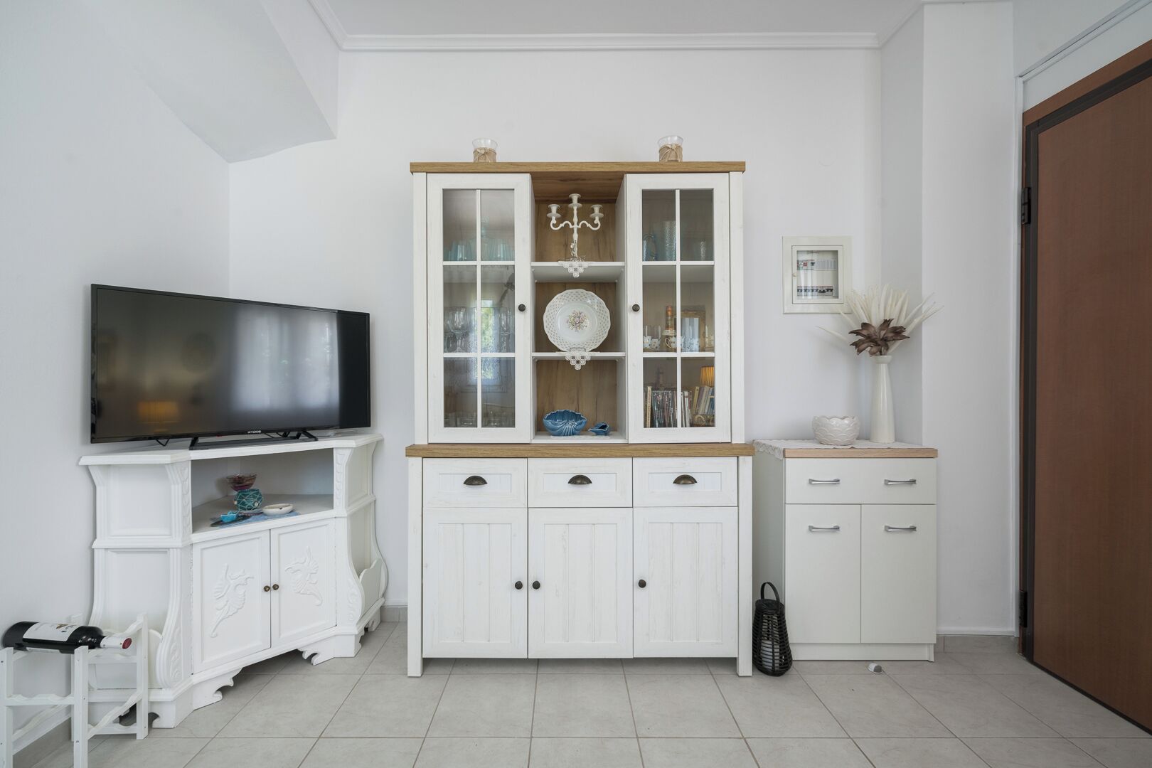 A room with a television on a white cabinet, a wooden cabinet with glass doors, and a small white cabinet with drawers.