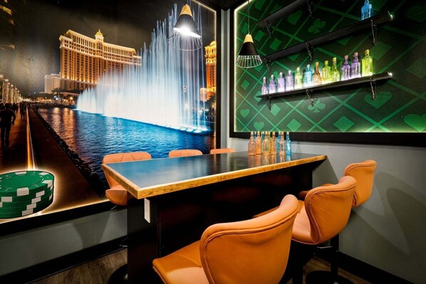 Un espace bar comprenant une table, des chaises, un mur orné d’un motif losangique vert et une étagère avec des bouteilles.