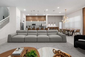 Un salon moderne avec un canapé d’angle gris, un coin repas avec des chaises en bois et une cuisine en arrière-plan.