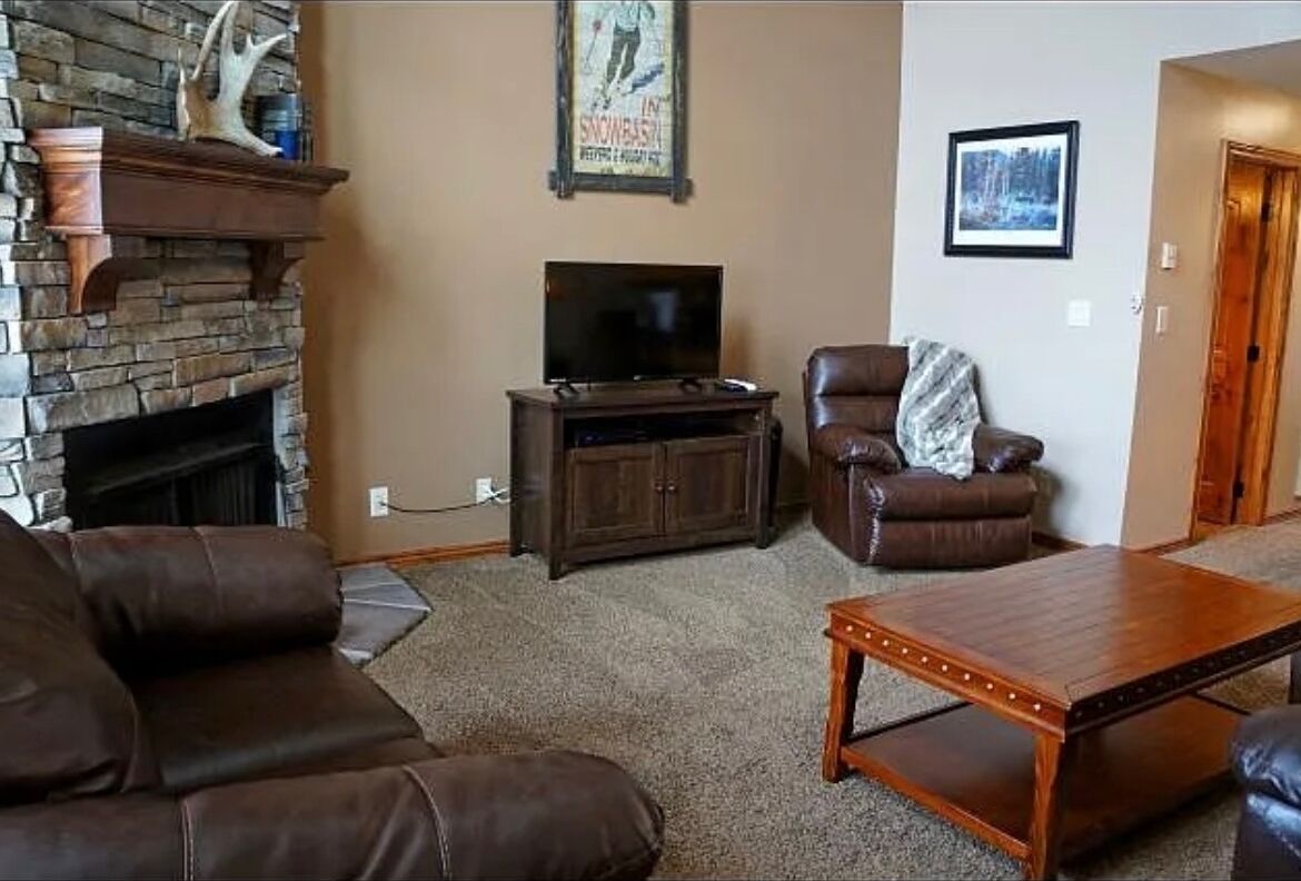 A living room with a stone fireplace, a television on a wooden stand, a leather armchair, and a coffee table.