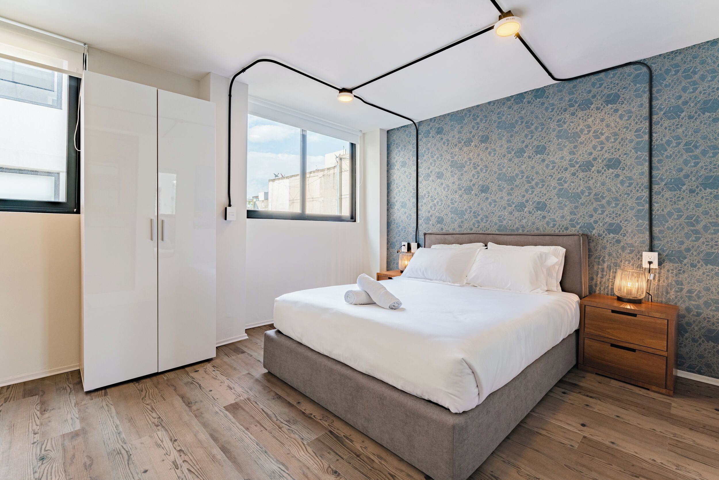 Ein modernes Schlafzimmer mit einem großen Bett, einem Nachttisch aus Holz, einer Wandlampe und einem Fenster mit Blick auf Gebäude.
