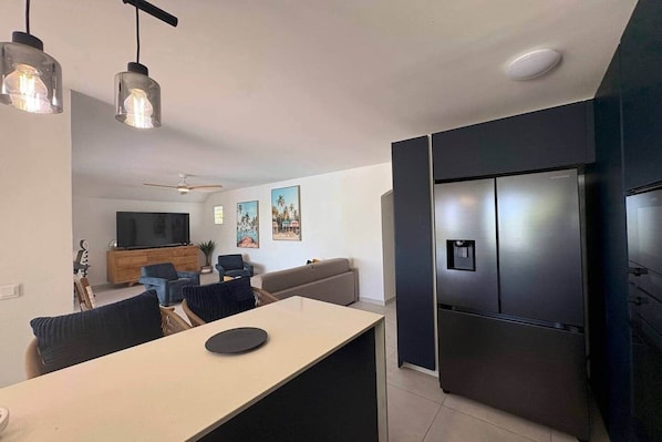 Uma cozinha moderna com frigorífico embutido, zona de refeições com mesa e cadeiras, e sala de estar com sofá e televisão.