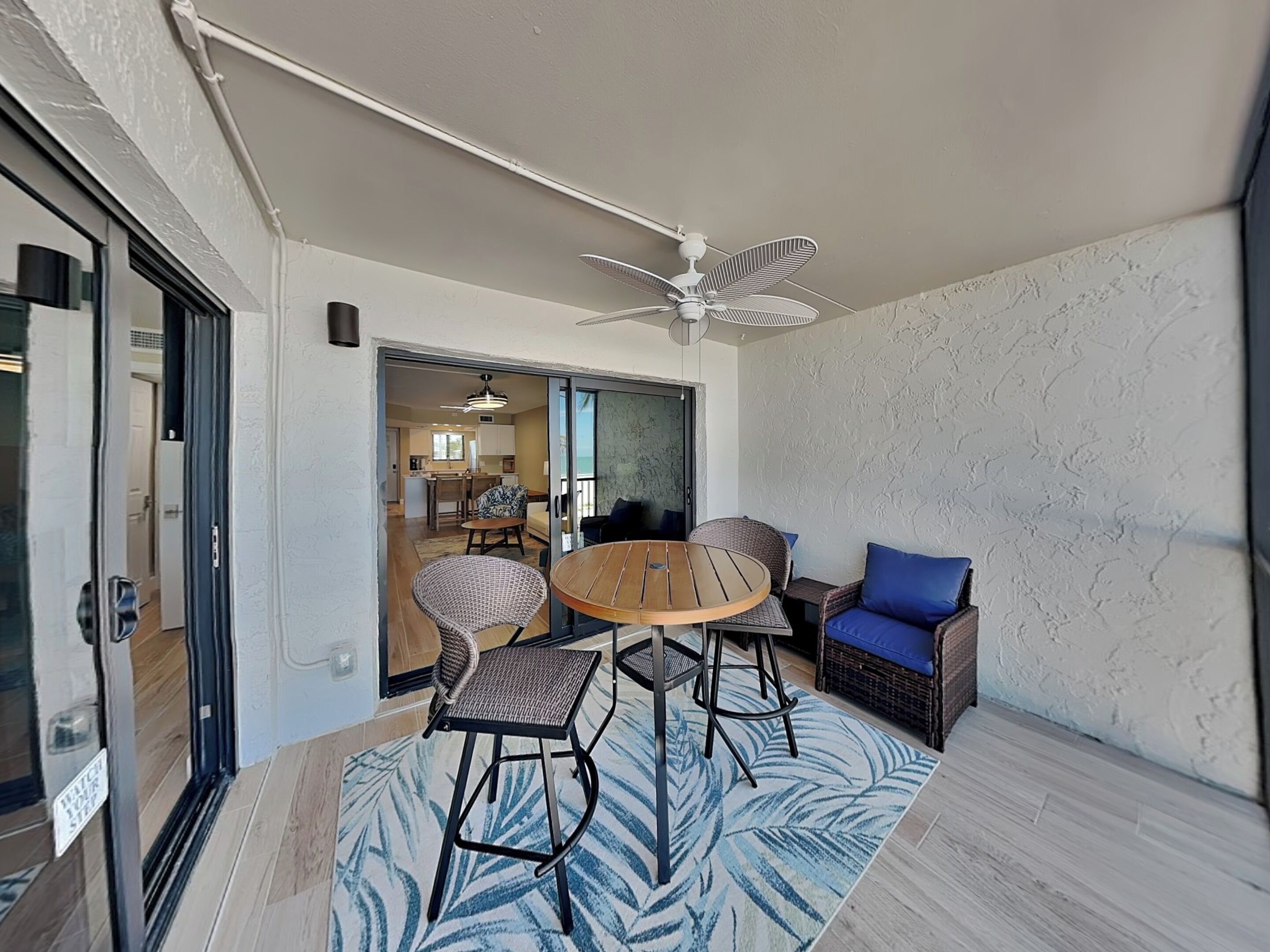 Une pièce dotée d’un ventilateur de plafond, d’une table à manger avec des chaises, d’un canapé en osier avec des coussins bleus et donnant sur la cuisine.
