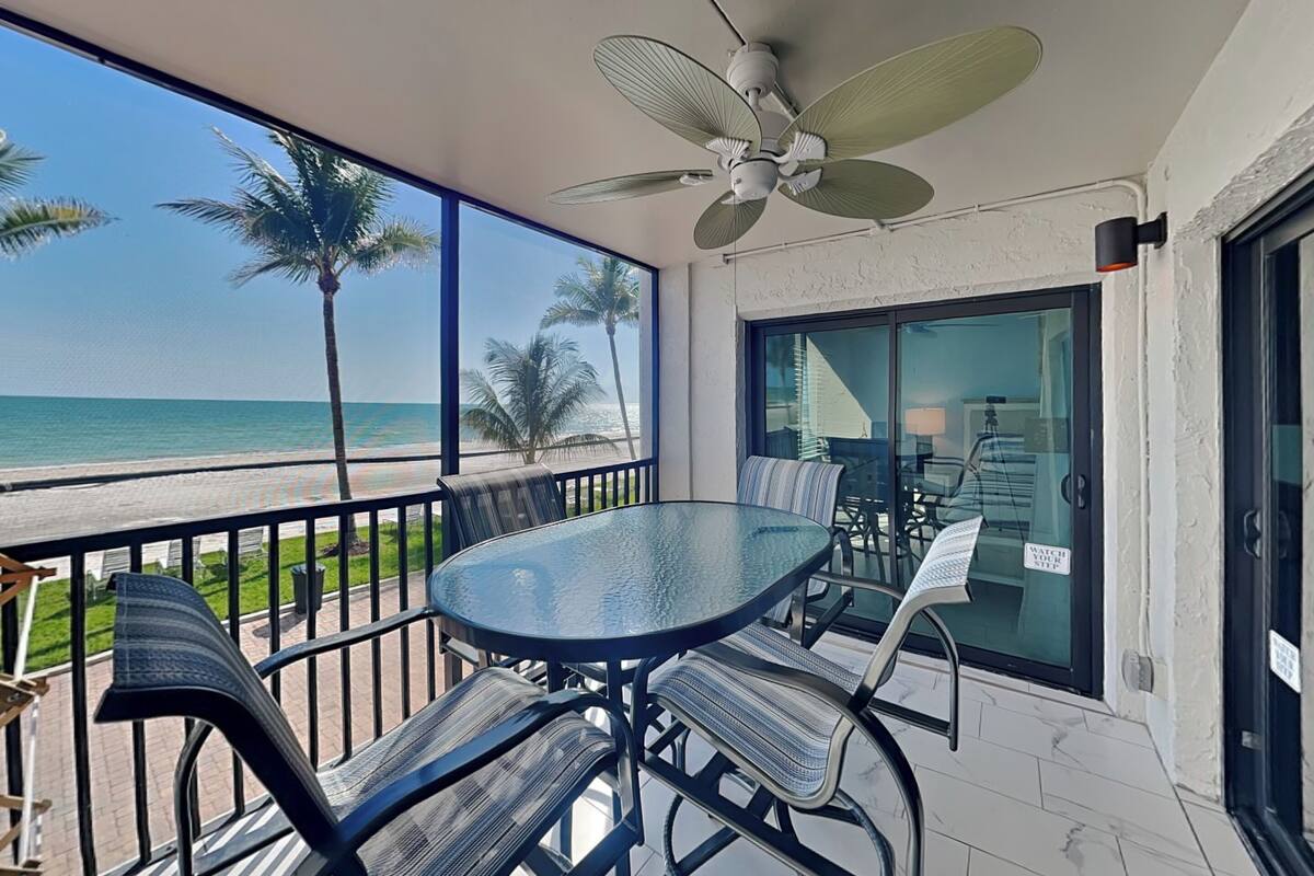 Un balcone con tavolo e sedie in vetro, una ventola a soffitto e vista sulla spiaggia e sull'oceano.